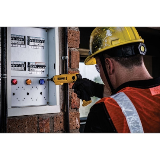 A worker wearing a yellow hard hat and orange safety vest uses a yellow DEWALT electric screwdriver to work on an electrical panel mounted on a brick wall.