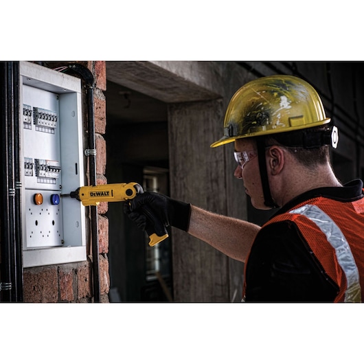 A person wearing a yellow safety helmet and orange reflective vest uses a yellow DEWALT cordless screwdriver (model DCF008) to work on an electrical panel mounted on a brick wall.