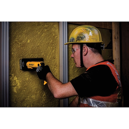 A worker wearing a yellow hard hat, orange safety vest, and black gloves uses a DEWALT DCF008 tool to install or prepare a section in a yellow textured wall on a construction site. The worker's face is blurred for privacy.
