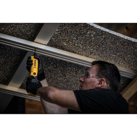 A person wearing black gloves uses a yellow DEWALT DCF008 tool to screw into a metal frame attached to a textured ceiling. The person's face is blurred for privacy.