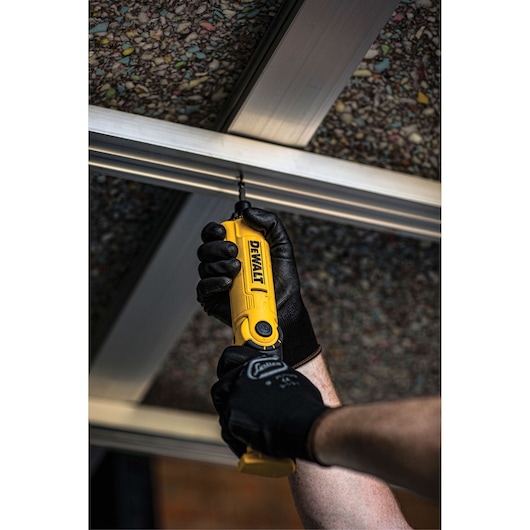 A person wearing black work gloves is using a yellow DEWALT cordless screwdriver to drive a screw into a metal frame, viewed from below.