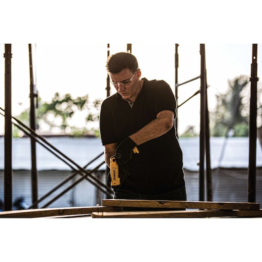 A person wearing a black shirt and black gloves is using a yellow DEWALT power tool to work on wooden planks at a construction site. The person's face is blurred. There are scaffolding structures and trees in the background.