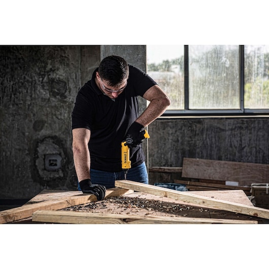 A person wearing black gloves is using a yellow DEWALT cordless drill to work on wooden planks in a workshop with a concrete wall and large window in the background. The person's face is obscured.