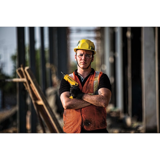 A person wearing a yellow hard hat and orange safety vest stands at a construction site, holding a yellow DEWALT power tool with crossed arms. The face is blurred for privacy.