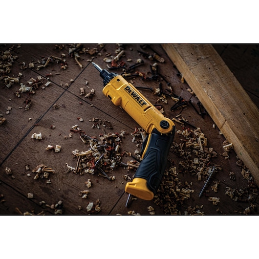 A yellow and black DEWALT cordless screwdriver (SKU: DCF008) lies on a wooden floor, surrounded by wood shavings and screws, with a piece of lumber nearby.