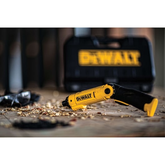 A yellow and black DEWALT electric screwdriver is lying on a wooden surface with wood shavings. A black DEWALT carrying case and safety glasses are in the background.