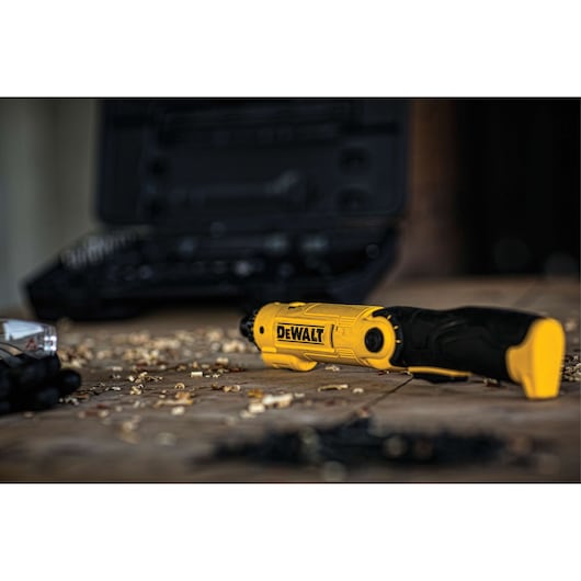 A DEWALT yellow and black electric screwdriver laying on a wooden surface with wood shavings and a black tool case in the background.