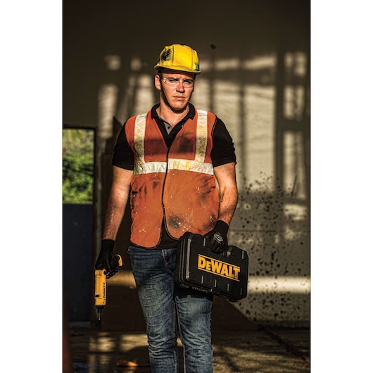 A construction worker wearing a yellow hard hat, orange safety vest, and gloves stands indoors holding a DEWALT tool case in one hand and a yellow DEWALT power tool in the other hand.