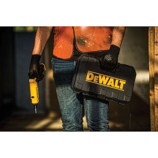 A person wearing an orange safety vest and gloves holds a yellow DEWALT power tool in one hand and a black DEWALT carrying case in the other hand.