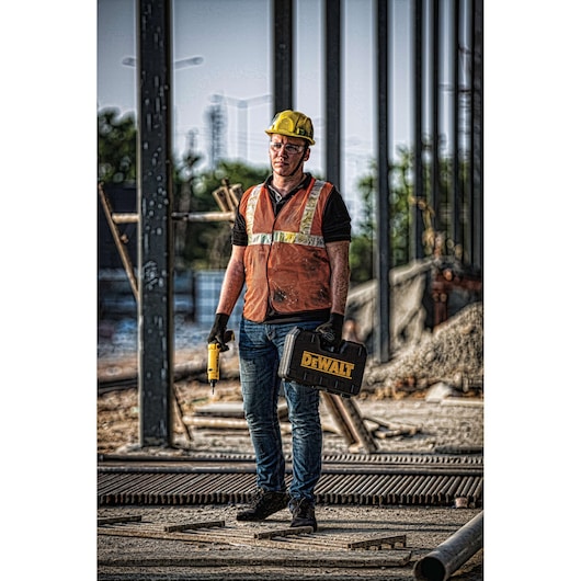 A construction worker wearing a yellow hard hat, orange safety vest, and gloves stands on a building site holding a DEWALT toolbox and a yellow cordless drill. The worker's face is blurred, and steel frameworks are visible in the background.