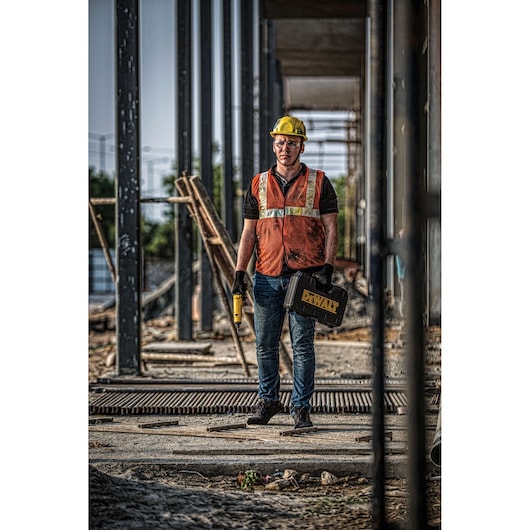 A construction worker wearing a yellow hard hat and an orange safety vest stands at a building site holding a DEWALT tool case and a cordless power tool. The worker is surrounded by steel structures and construction materials.