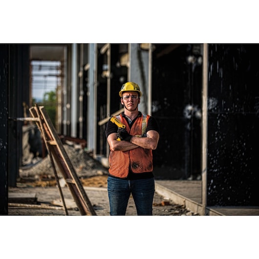 A construction worker wearing a yellow hard hat and orange safety vest stands with arms crossed at a construction site, holding a yellow DEWALT power tool. The face is obscured for privacy.