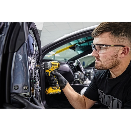 A person wearing black gloves uses a yellow DEWALT cordless screwdriver (DCF601D2) to work on the interior of a car door. The individual's face is blurred, and they are wearing a DEWALT-branded shirt.