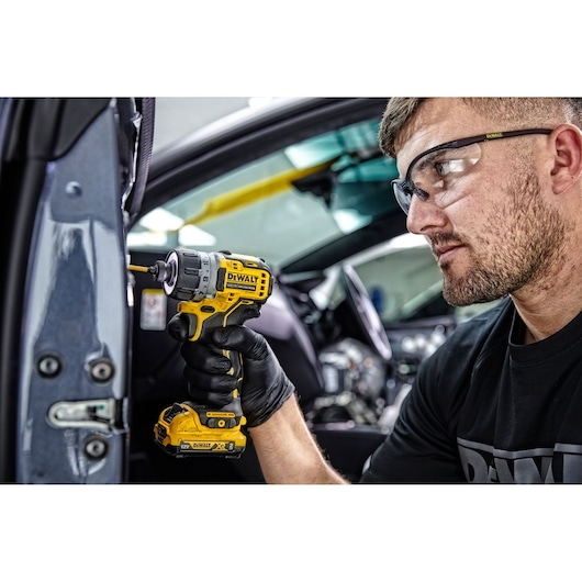A person wearing black gloves uses a yellow DEWALT DCF601D2 cordless screwdriver to work on a part inside a vehicle. The person's face is blurred for privacy.