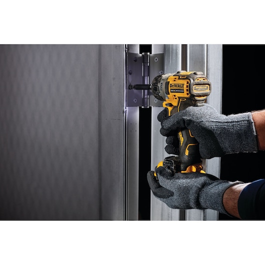 Gloved hands use a DEWALT drill to install a hinge on a metal door frame.