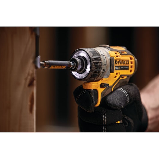A DEWALT cordless drill being used to drive a screw into wood by a person wearing black work gloves.