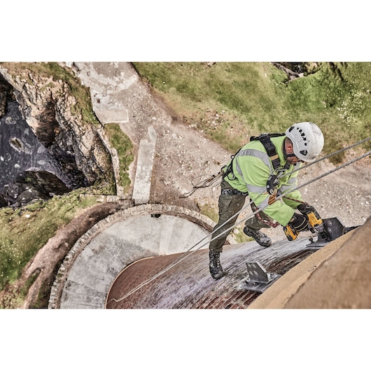 Worker uses DEWALT tool on harnessed climb, repairing structure outdoors above stone and grass area.