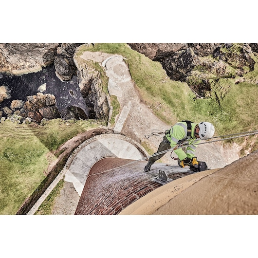 Worker with safety gear drills on a high brick tower near rocky cliffs and grass.