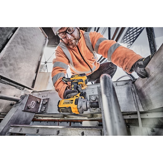 Worker uses DEWALT DCF894P2 impact wrench on metal structure at construction site, wearing safety gear.