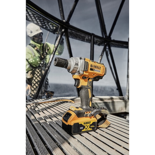 DEWALT DCF894P2 impact wrench with battery on metal floor, outdoor worker blurred in background.