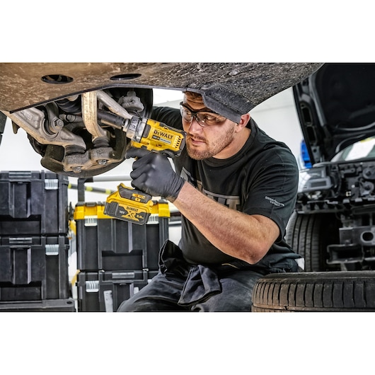 Person uses DEWALT DCF894P2 impact wrench on car in workshop. Toolboxes and tire are visible.