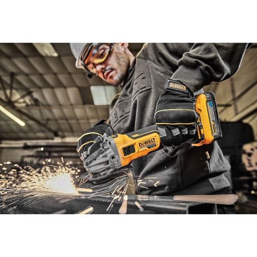 DEWALT cordless grinder in use, cutting metal with sparks, worker wearing gloves and safety gear in workshop.