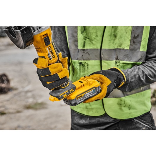 Worker holds DEWALT DCG405H2T grinder with yellow gloves and a green safety vest at a construction site.