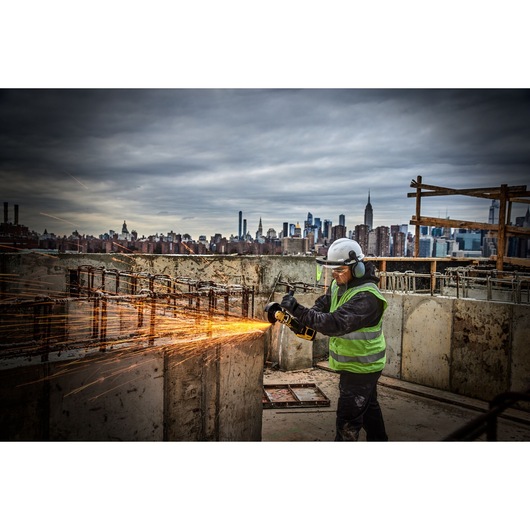 Worker using DEWALT grinder on construction site, sparks flying, city skyline in background.