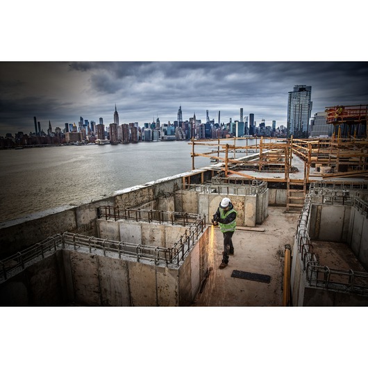 Worker uses power tool on concrete at construction site near waterfront, city skyline in background.
