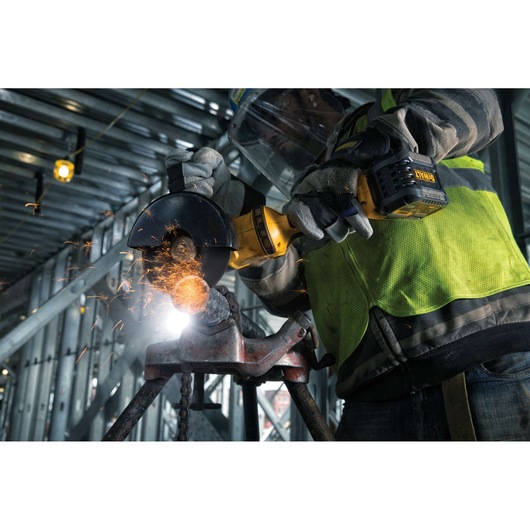 A worker using a DEWALT cordless angle grinder (model DCG414T2) to cut metal, generating sparks. The worker is wearing safety gear including gloves, a visor, and a reflective vest, in an industrial setting.
