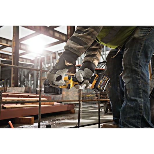 A person wearing gloves and safety gear uses a DEWALT cordless angle grinder to cut through metal rods at a construction site. Sparks are flying from the metal as the tool operates.