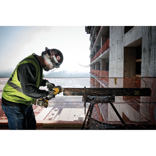A construction worker wearing safety gear and a helmet uses a DEWALT cordless angle grinder to cut through a metal pipe. Sparks are flying as the tool makes contact with the pipe. The scene takes place at an outdoor construction site with an unfinished building in the background.
