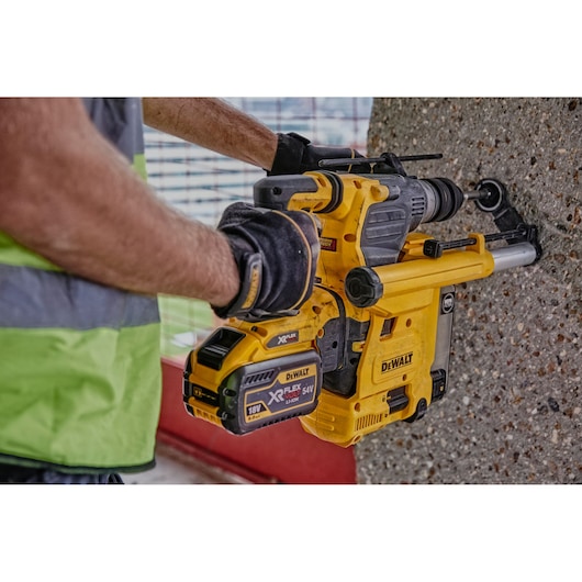 A person wearing work gloves and a safety vest is using a DEWALT cordless rotary hammer drill to bore into a concrete wall.