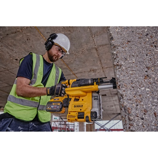 A person wearing safety gear and a high-visibility vest operates a yellow DEWALT DCH334X2 rotary hammer drill against a concrete wall at a construction site.