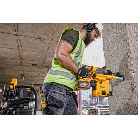 Construction worker wearing a safety vest, helmet, and ear protection using a yellow DEWALT cordless rotary hammer drill on a concrete wall at a building site.