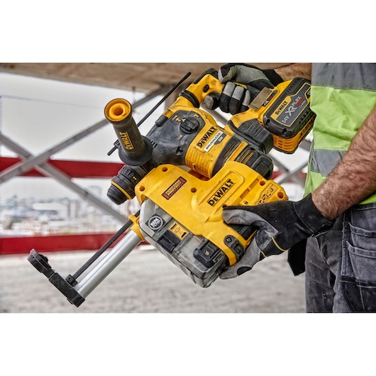 A person wearing gloves and a safety vest is holding a DEWALT cordless rotary hammer drill, model DCH334X2, at a construction site.