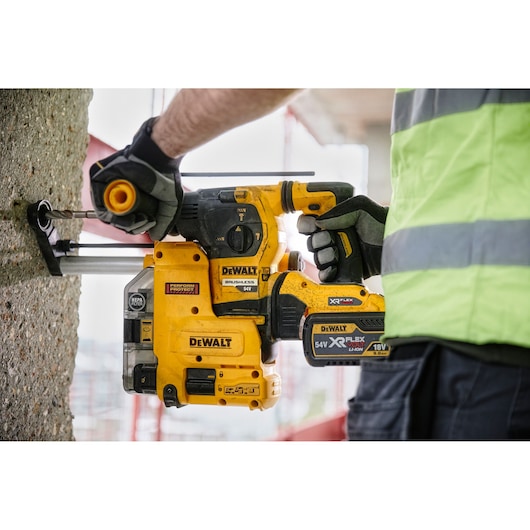 A person wearing gloves and a high-visibility vest is using a yellow DEWALT DCH334X2 cordless rotary hammer drill with a brushless motor to drill into a concrete wall.