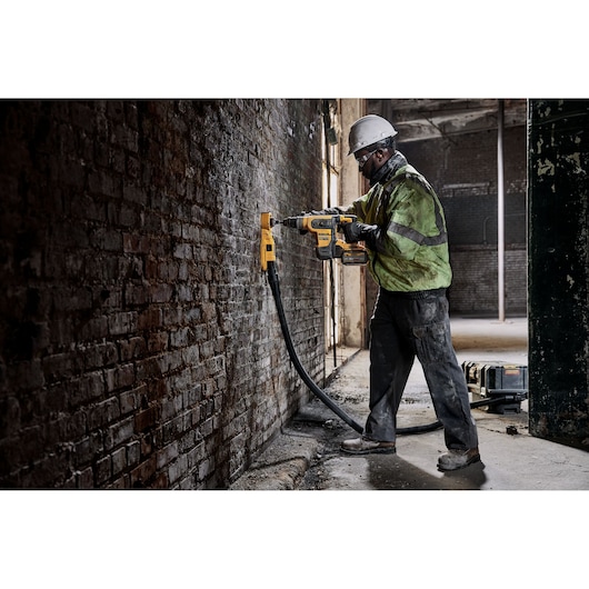Worker uses DEWALT rotary hammer drill on brick wall, wearing helmet and safety jacket in industrial setting.