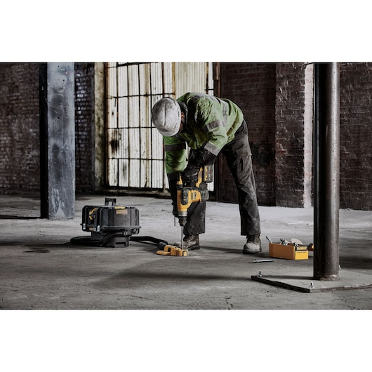 Worker drills concrete floor using DEWALT power tool; toolbox and equipment visible nearby in industrial setting.