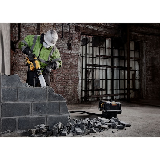Worker uses DEWALT power tool to demolish concrete wall in an industrial setting with rubble on the ground.