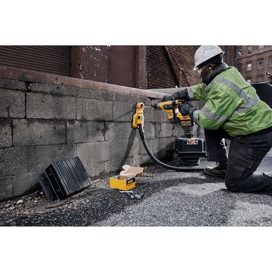Worker uses DEWALT rotary hammer drill DCH417X2 on a brick wall at a construction site; tools and parts nearby.