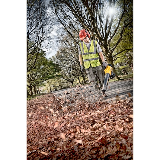 Person using a DEWALT blower to clear leaves on a park path, wearing high-vis vest and helmet.
