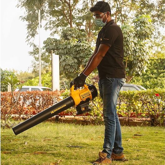 Person using a DEWALT DCM562P1 cordless leaf blower outdoors on grass near bushes.