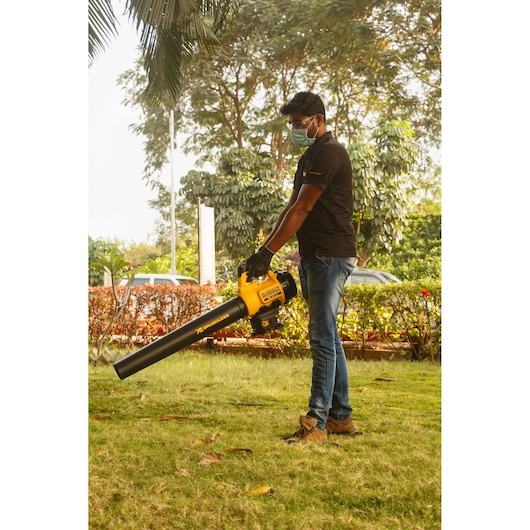 Person using a DEWALT DCM562P1 leaf blower outdoors on a grassy lawn with trees in the background.