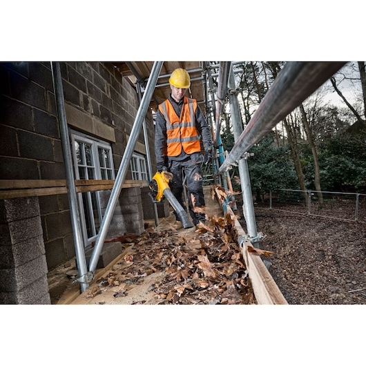 Worker uses a DEWALT leaf blower to clear leaves from scaffolding at a construction site.