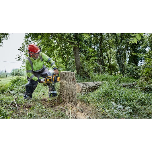 A person wearing safety gear uses a DEWALT cordless chainsaw to cut through a large tree stump outdoors, surrounded by grass and trees.