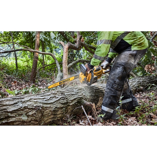 A person wearing protective gear uses a DEWALT cordless chainsaw to cut through a large fallen tree in a forested area.