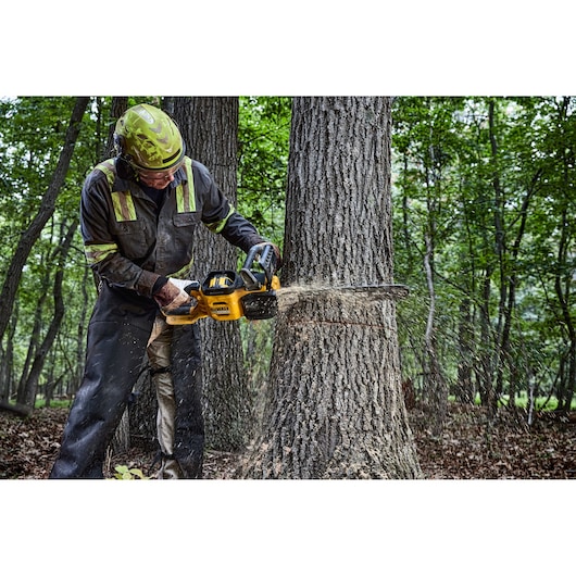 A person wearing protective gear operates a yellow DEWALT chainsaw while cutting into a tree trunk in a forest setting. Sawdust is visible as the chainsaw cuts through the wood.