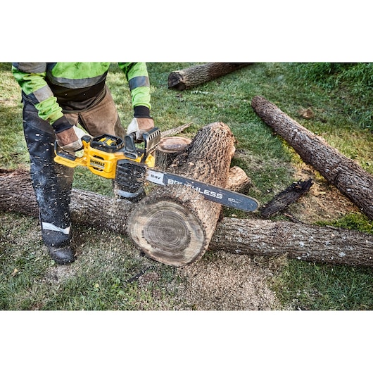 A person using a yellow DEWALT cordless chainsaw to cut through a large log outdoors. The chainsaw is in the process of cutting through the wood, producing wood shavings.