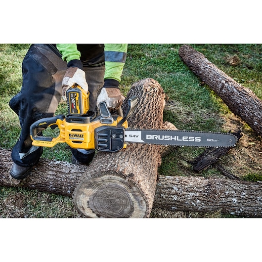A person wearing protective gloves and gear uses a DEWALT 54V brushless cordless chainsaw to cut a large tree log outdoors.